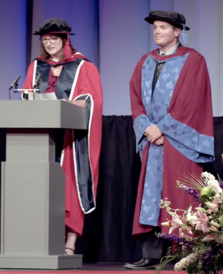 Graduation ceremony, two figures in red and blue robes on stage at the University of Brighton