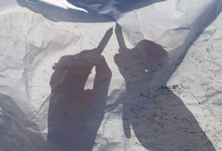 Artist Jane Fox's hands visible in silhouette and shadow through a crinkled white cloth making marks on the wind-blown material