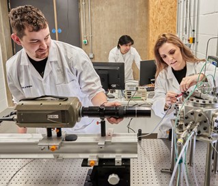 Three researchers in white coats work within a workshop environment on engineering research with mechanical objects and tools