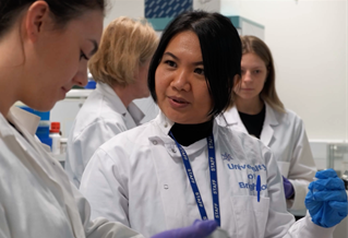 Post-doctoral chemistry researcher Dr Wulan Koagouw in laboratory with students. Scientists in white coats with blue gloves and equipment.