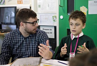 Male primary teacher sits at desk talking to pupil