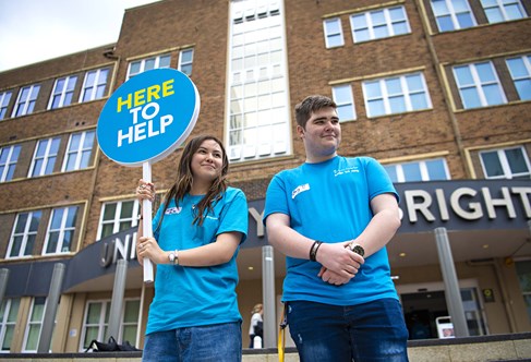 Two open day helpers in blue T-shirts holding a sign that reads 'Here to help'