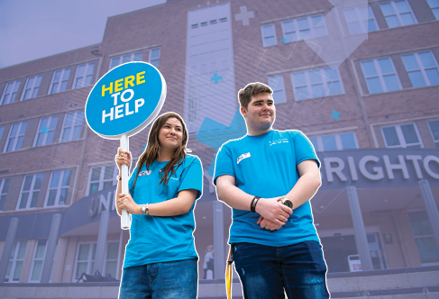 Two student ambassadors at an open day holding a 'Here to help' sign